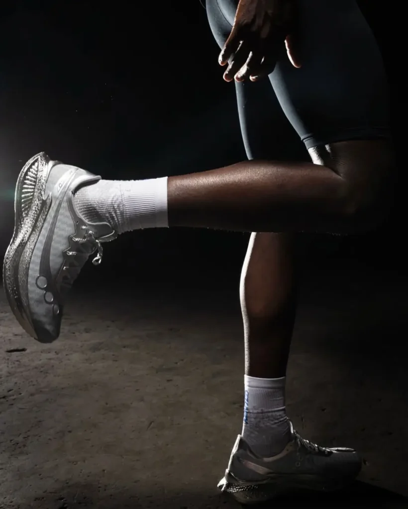 A runner is captured mid-stretch in a low-lit studio setting, one leg extended forward while the other supports their stance. They wear fitted dark athletic shorts, white crew socks, and sleek metallic running shoes with a sculpted, performance-driven sole. Dramatic side lighting highlights the contours of the leg muscles and the shoe’s reflective detailing, while the dark background isolates the movement and form, giving the scene a focused, almost cinematic intensity