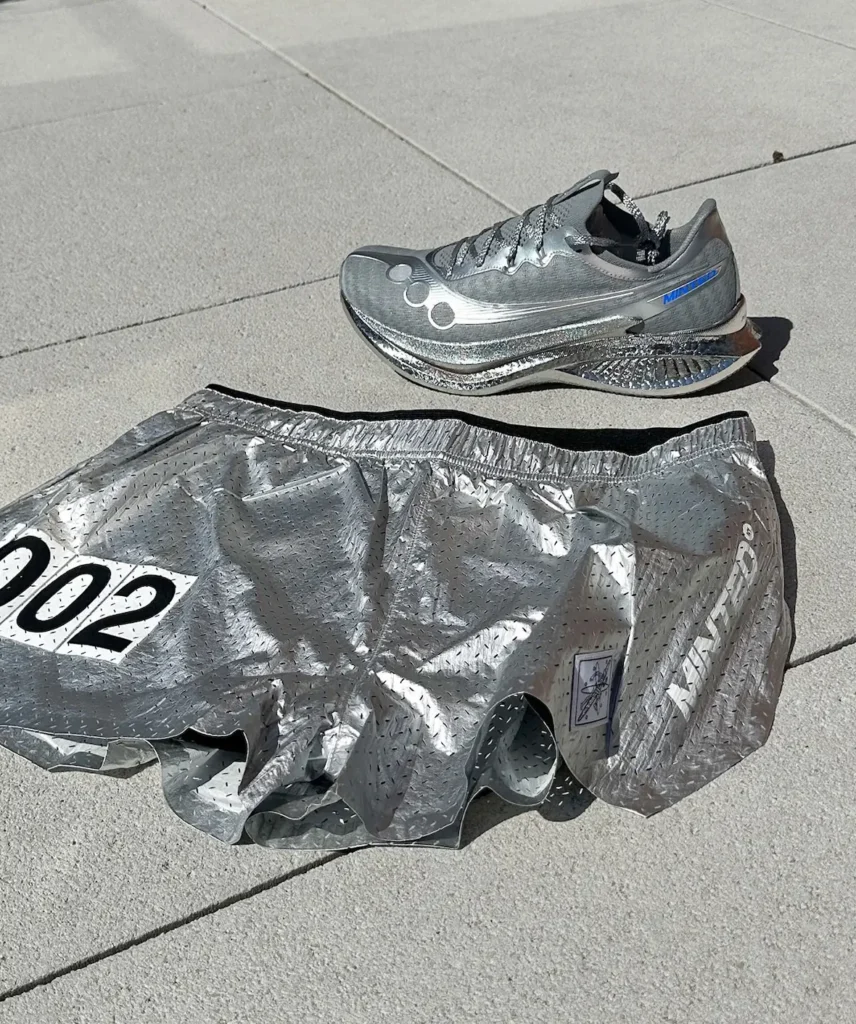 A metallic silver running shoe and matching lightweight shorts are laid out on a sunlit concrete surface. The shoe features a sleek, aerodynamic silhouette with subtle blue accents and a sculpted midsole, while the shorts have a crinkled, reflective texture with perforations, an elastic waistband, and bold printed detailing including the number “002"