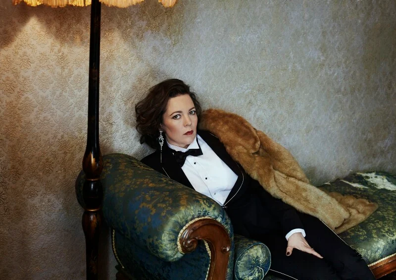 Olivia Colman reclines across a vintage chaise lounge in a sharply tailored tuxedo and draped fur stole, embodying the theatrical elegance and emotional complexity associated with Isabella Blow in Wild Bird. Framed by warm ambient lighting and richly textured interiors, the portrait channels aristocratic glamour with an undercurrent of melancholy, reflecting the film’s exploration of fashion, identity, and creative vulnerability