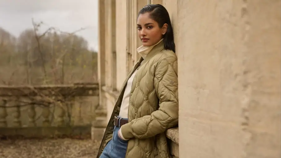 Model leaning against a stone exterior wall while wearing an olive quilted Bench jacket layered over a cream high-neck top and blue denim. The muted outdoor setting and soft neutral tones create a calm, understated mood that highlights the brand’s functional everyday outerwear aesthetic
