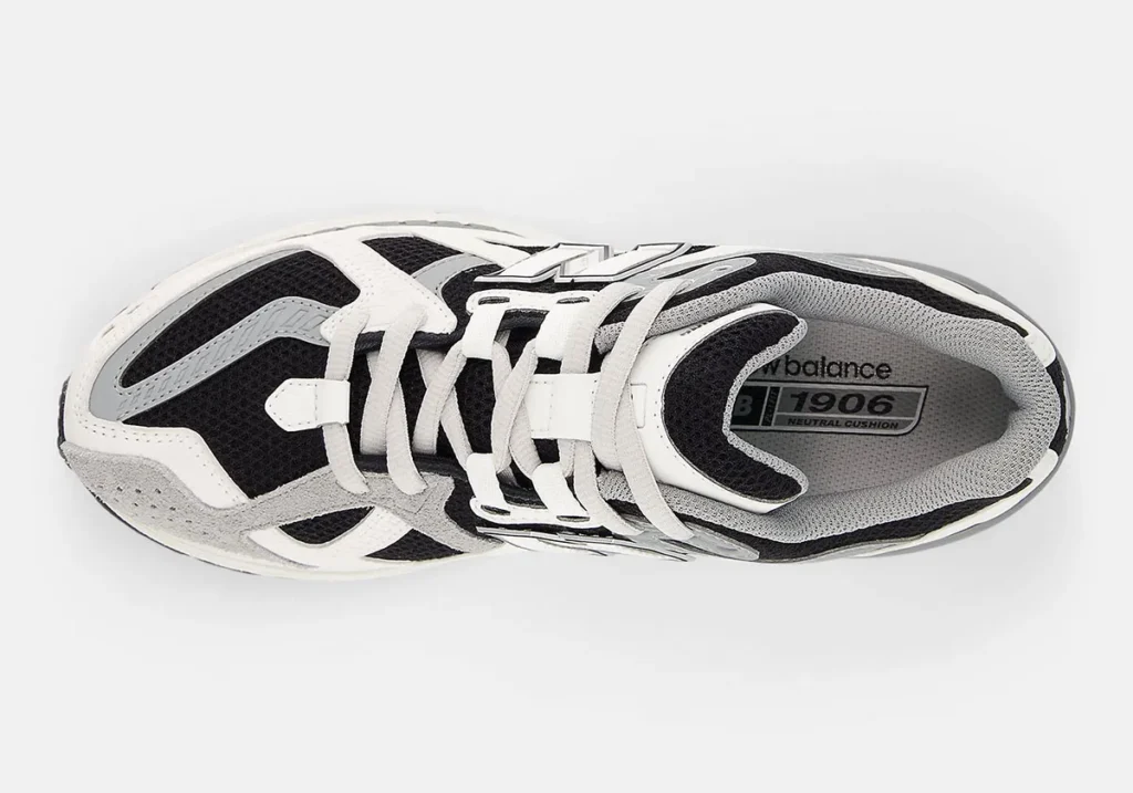 Side profile view of the New Balance 1906 shoe in a black, white, and grey palette featuring breathable black mesh underlays, layered white synthetic overlays, grey suede detailing around the toe, and a large outlined “N” logo at the midfoot. The retro-inspired runner silhouette is paired with sculpted cushioning, textured midsoles, and metallic grey heel support components that emphasize the shoe’s early-2000s performance-running aesthetic and technical streetwear appeal