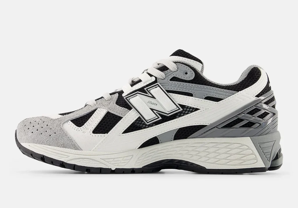 Side profile view of the New Balance 1906 shoe in a black, white, and grey palette featuring breathable black mesh underlays, layered white synthetic overlays, grey suede detailing around the toe, and a large outlined “N” logo at the midfoot. The retro-inspired runner silhouette is paired with sculpted cushioning, textured midsoles, and metallic grey heel support components that emphasize the shoe’s early-2000s performance-running aesthetic and technical streetwear appeal