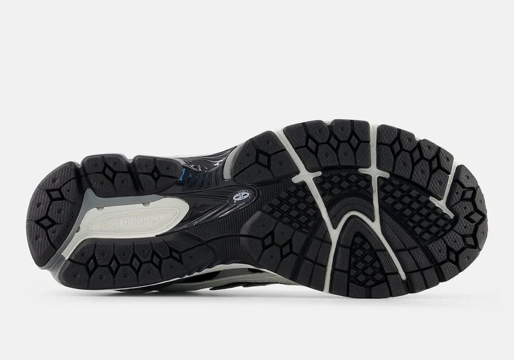 Outsole view of the New Balance 1906 showcasing a heavily segmented black rubber traction pattern designed for stability, flexibility, and impact control. The sole features geometric tread pods across the forefoot and heel, separated by white flex grooves that enhance movement and cushioning responsiveness. A sculpted midfoot support shank and visible Stability Web detailing reinforce the shoe performance-running foundation, while subtle grey and off-white accents break up the otherwise dark outsole composition. The aggressive tread layout and layered tooling emphasize the shoe’s technical early-2000s runner aesthetic and durability-focused construction
