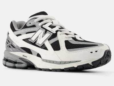 Front-angled view of the New Balance 1906 in a black, white, and grey colorway highlighting the shoe layered retro-runner construction with breathable black mesh, white synthetic overlays, and metallic grey detailing around the heel and eyestays. The oversized outlined “N” logo stands prominently along the sidewall, while sculpted ABZORB cushioning pods and segmented outsole tooling emphasize the model’s technical performance heritage. Curved paneling, textured midsoles, and layered materials give the silhouette a dynamic early-2000s running aesthetic balanced with contemporary streetwear style