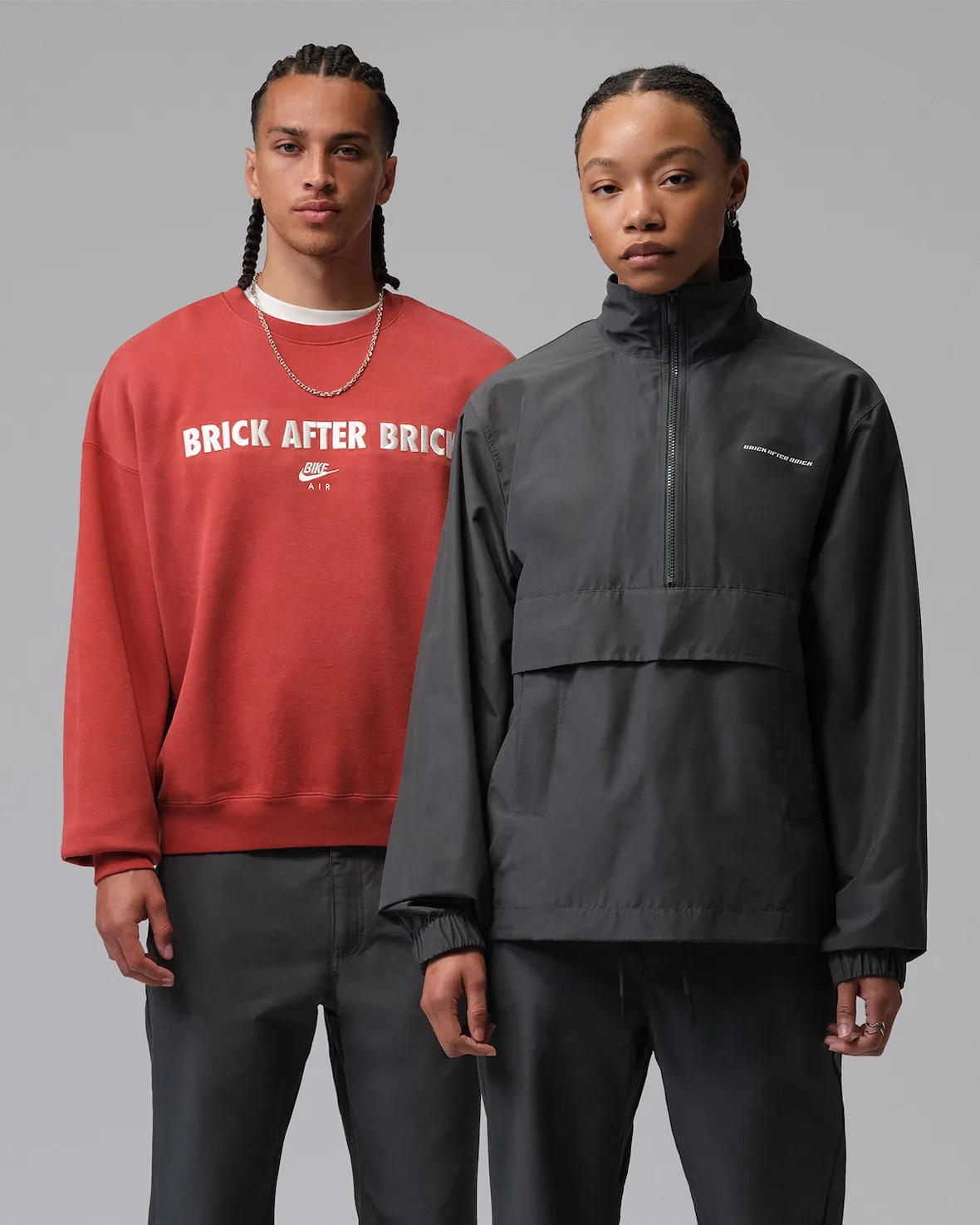 A studio campaign image featuring two models showcasing pieces from the Nigel Sylvester x Jordan Brand “Brick After Brick” apparel collection against a clean gray backdrop. One model wears a washed red crewneck sweatshirt emblazoned with oversized “BRICK AFTER BRICK” lettering across the chest alongside co-branded Bike Air graphics, while the other appears in a minimalist charcoal half-zip pullover jacket with subtle tonal branding near the chest. Both looks are paired with relaxed dark trousers, emphasizing the collection’s understated utilitarian styling and streetwear-driven silhouette balance. The image reflects Nigel Sylvester’s BMX-rooted aesthetic through muted industrial tones, oversized fits, and construction-inspired branding language that complements the accompanying Air Jordan 4 collab