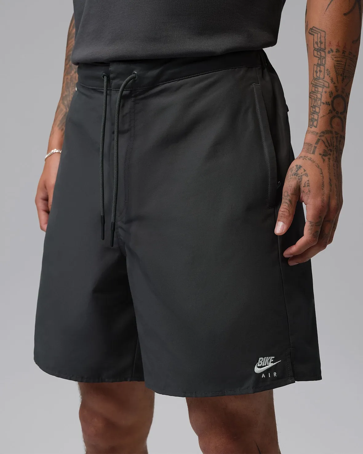 A close-up studio image highlighting a pair of minimalist black shorts from the Nigel Sylvester x Jordan Brand “Brick After Brick” apparel collection. The shorts feature a lightweight technical fabric construction with a relaxed fit, elastic waistband, and extended tonal drawstrings that reinforce the collection’s utilitarian aesthetic. Near the hem, co-branded “Bike Air” graphics appear in white embroidery, referencing Nigel Sylvester’s BMX culture influence and reinterpretation of classic Nike Air iconography. The understated design, paired with clean tailoring and muted industrial tones, aligns with the collection’s functional streetwear direction and complements the accompanying Air Jordan 4 collab