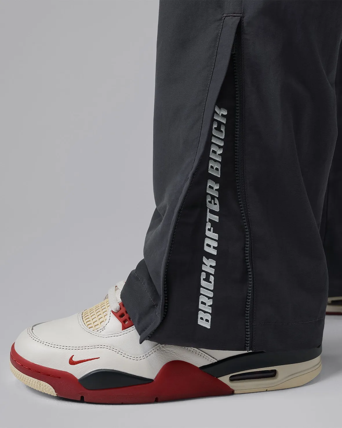 A detailed styling image from the Nigel Sylvester x Jordan Brand “Brick After Brick” apparel collection, focusing on the lower-leg construction of a pair of technical black pants worn over the collaborative Air Jordan 4 sneakers. The pants feature elongated side zippers that open to reveal bold vertical “BRICK AFTER BRICK” typography in white, introducing a functional BMX-inspired design element with customizable ventilation and movement. Beneath the pants, the Air Jordan 4 appears in off-white, red, cream, and dark navy tones, highlighting the sneaker’s signature mesh netting, embroidered mini Swoosh, and sculpted midsole with visible Air cushioning. The composition emphasizes the coordinated relationship between the footwear and apparel capsule, blending utilitarian sportswear detailing with Nigel Sylvester’s industrial streetwear aesthetic