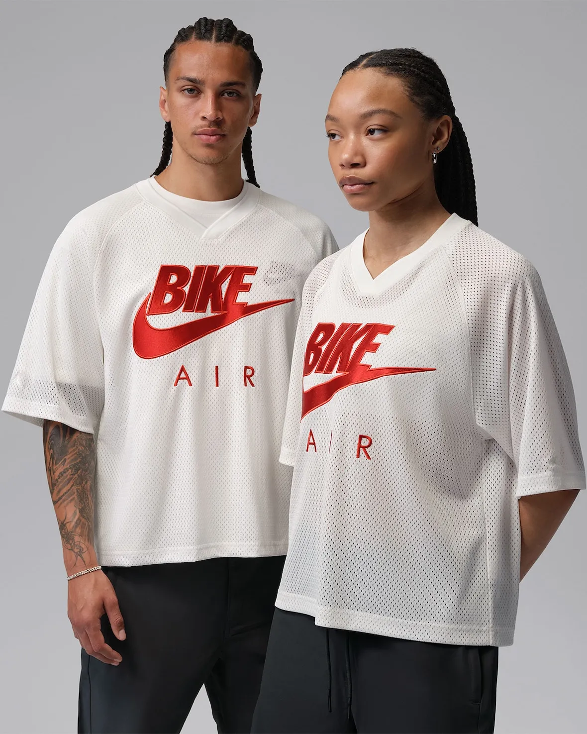 A studio campaign image featuring two models wearing oversized mesh jerseys from the Nigel Sylvester x Jordan Brand “Brick After Brick” apparel collection against a neutral gray backdrop. The tops arrive in an off-white perforated athletic fabric with large red “Bike Air” graphics stretched prominently across the chest, reimagining classic Nike Air branding through Nigel Sylvester’s BMX-inspired lens. The relaxed silhouettes, dropped shoulders, and breathable sportswear construction give the pieces a retro practice-jersey feel while maintaining a contemporary streetwear proportion. Paired with loose black trousers, the styling reinforces the collection’s fusion of athletic heritage, industrial minimalism, and BMX culture aesthetics that accompany the collab Air Jordan 4 release