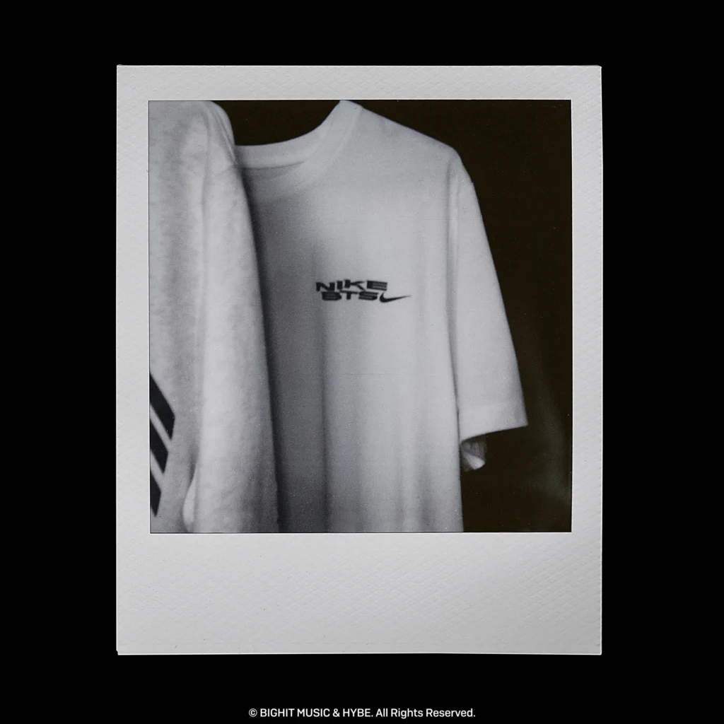 Minimalist black-and-white image of a Nike BTS T-shirt with a small chest logo hanging beside a graphic hoodie