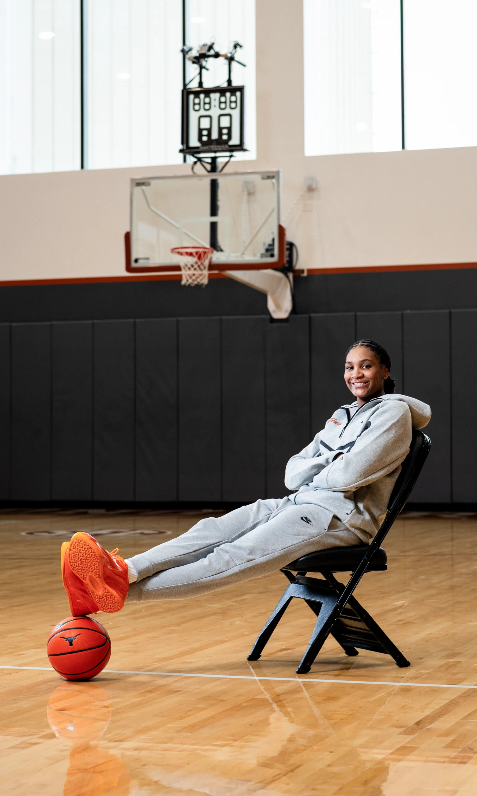 Madison Booker relaxes courtside inside Texas’ practice gym, dressed in a grey Nike tracksuit with bright orange KD sneakers balanced on a basketball beneath her feet, framed by the quiet atmosphere of the empty court behind her