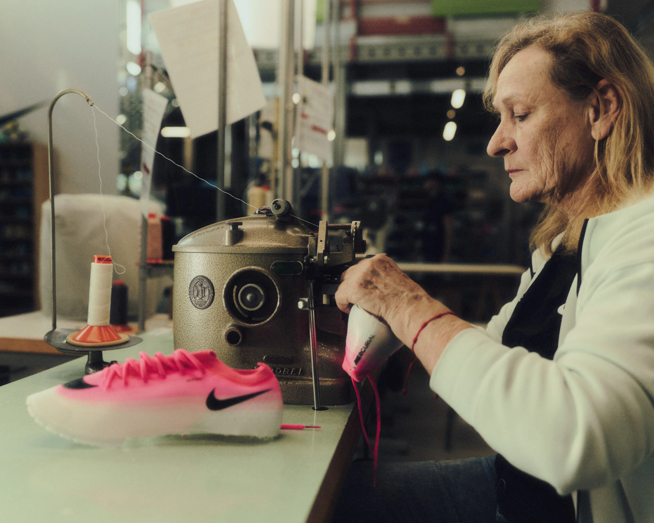 Italian craftsperson hand-finishing a vivid pink Nike Mercurial football boot beside an industrial sewing machine inside a specialist footwear workshop