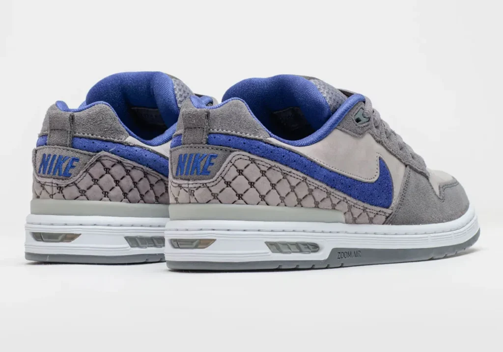 Rear three-quarter product view of a low-top Nike SB shoe shown as a pair in flint grey and deep royal blue. The design combines grey suede overlays with smooth grey panels, a textured royal blue Swoosh, and a monogram-style geometric print wrapping the heel. Blue embroidered Nike branding appears on the back, while a white midsole with view Zoom Air cushioning reinforces the skate-focused construction