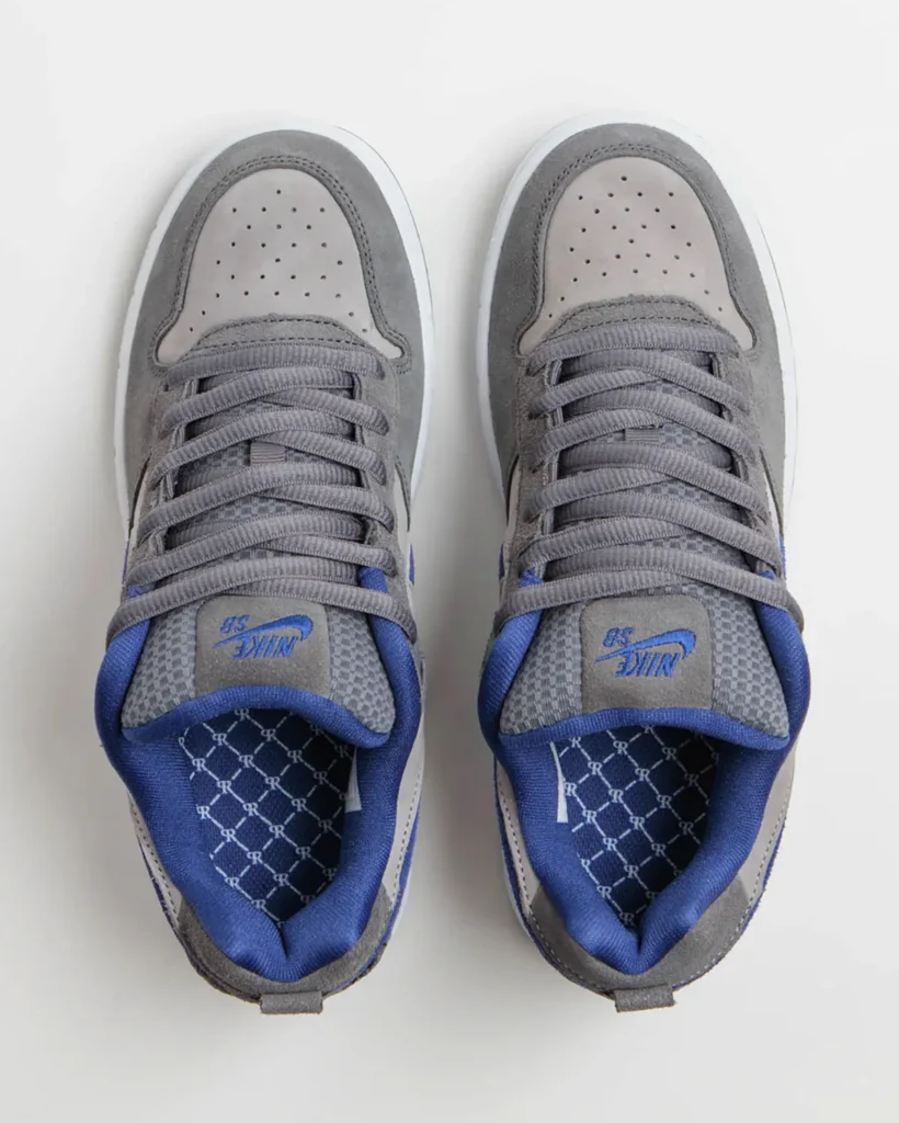 Top-down product view of a pair of Nike SB low-top sneakers in flint grey and deep royal blue. The design highlights grey suede overlays, perforated light grey toe boxes, wide grey skate laces, textured mesh tongues with blue Nike SB branding, and royal blue padded lining. The interior footbeds feature an all-over monogram-style graphic pattern that complements the heel detail