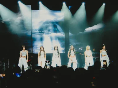 NMIXX performs on stage during a live concert, dressed in coordinated white outfits beneath bright spotlights, with a dramatic ocean-inspired visual backdrop illuminating the venue as fans record the performance on their phone