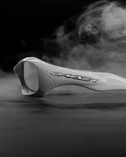 Black-and-white close-up of white Oakley FatCat sunglasses photographed in atmospheric smoke, emphasizing the elongated wraparound profile, sculpted frame contours, and oversized Oakley logo across the temple
