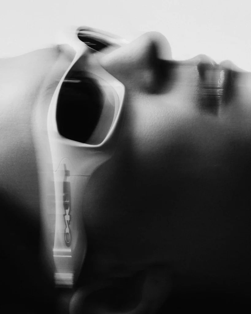Black-and-white editorial portrait of a reclining person wearing oversized white Oakley FatCat sunglasses, captured with soft motion blur and high-contrast lighting to emphasize the sculptural frame and futuristic Y2K aesthetic