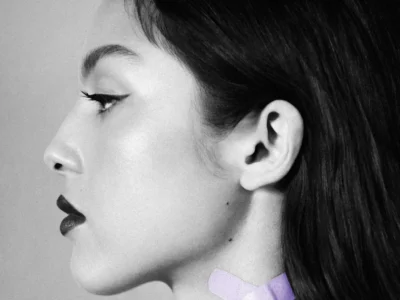 Black-and-white close-up side profile portrait of a woman facing left, photographed in soft studio lighting. She wears sharp winged eyeliner, dark lipstick, and has long dark hair pulled back behind the ear. The composition emphasizes clean facial contours, smooth skin texture, and a minimalist beauty-editorial aesthetic. A small pale lavender patch or graphic detail appears near the side of the neck, adding subtle contrast to the monochrome image