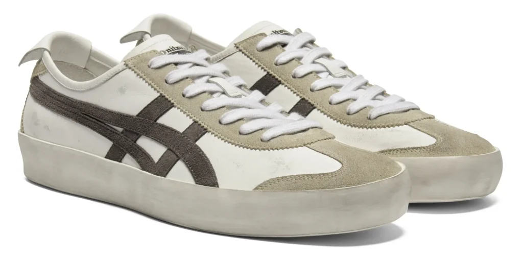 Pair of off-white and cream Onitsuka Tiger low-top sneakers shown in a clean studio product shot, featuring distressed leather panels, soft suede overlays, dark textured side stripes, and thick vintage-inspired rubber soles. The minimal neutral palette and worn-in finish emphasize handcrafted character and elevated Italian-made style