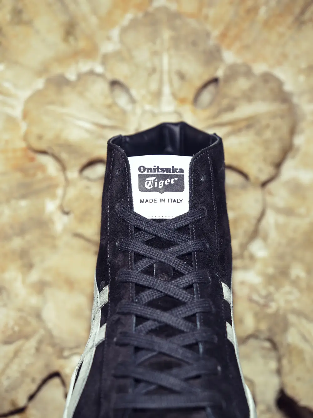 Front-facing close-up of a black Onitsuka Tiger high-top shoe featuring tonal laces and a white tongue label marked “Made in Italy.” Set against an aged stone backdrop with ornamental floral relief, the composition emphasizes premium suede texture, vintage-inspired detailing, and understated Italian craft