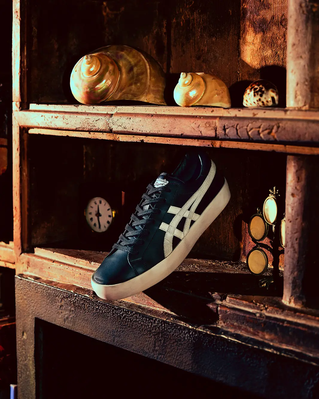Black and cream Onitsuka Tiger low-top shoe displayed within a weathered wooden cabinet surrounded by curated vintage objects, including seashells, a small clock, and brass-framed eyewear. Warm directional lighting highlights the smooth leather upper, contrast side stripes, and minimalist silhouette, creating a nostalgic still-life atmosphere with artisanal, collected appeal