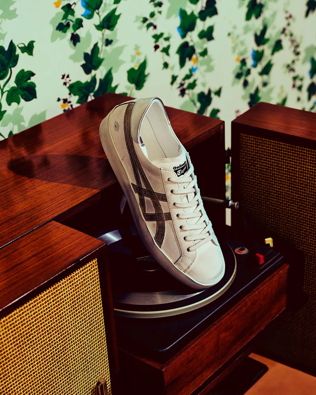 Cream and off-white low-top Onitsuka Tiger sneaker displayed diagonally atop a vintage wooden turntable setup, framed by rich walnut speakers and set against botanical ivy-pattern wallpaper. The scene blends retro audio aesthetics with understated luxury, highlighting textured side stripes, soft leather panels, and warm ambient lighting