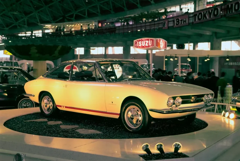 Early Isuzu 117 Coupé displayed at an auto show, featuring the original quad-round-headlight front end, Pininfarina-inspired elegance, and one of Japan’s most iconic classic coupe design