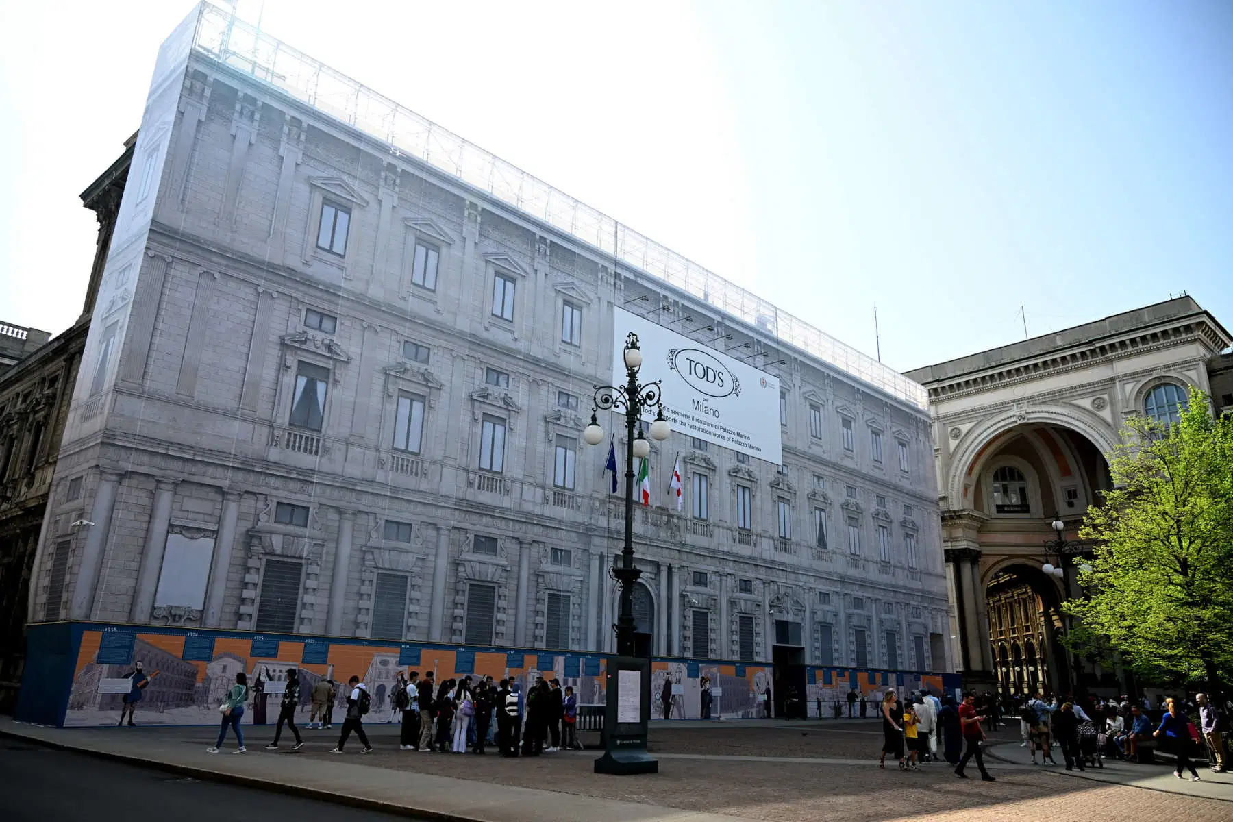 Exterior view of Palazzo Marino during its restoration phase in Piazza della Scala, wrapped in architectural scaffolding mesh bearing Tod’s Group branding, as pedestrians gather outside the historic Renaissance civic landmark beside the monumental arcade entrance of the Galleria area in central Milan