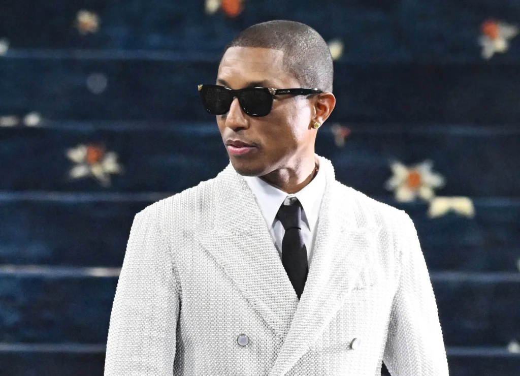 Pharrell Williams wearing a textured ivory double-breasted suit with black tie and sunglasses, standing against a dark patterned backdrop with subtle floral accents, projecting refined, minimalist elegance at a formal event