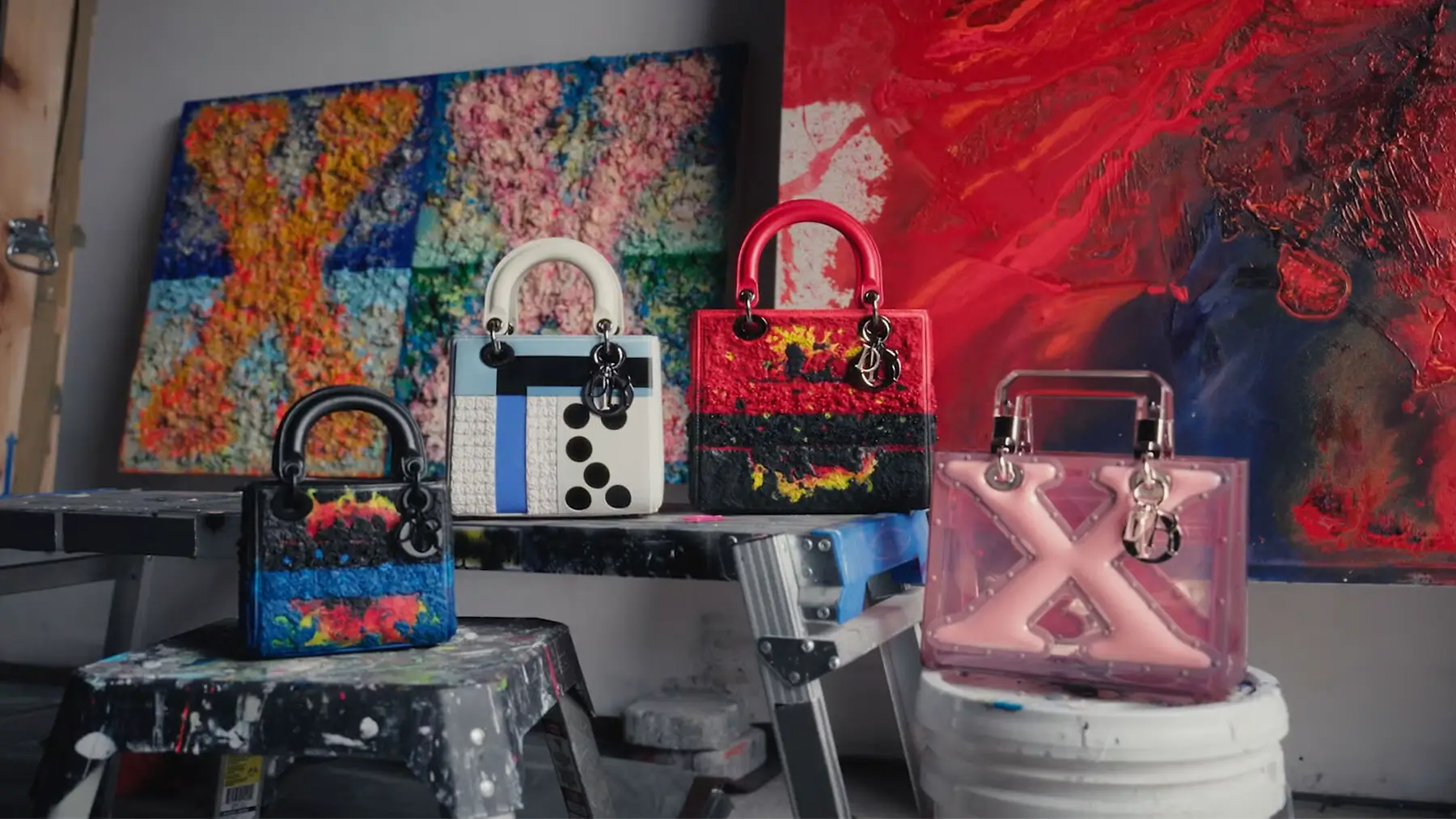 A curated display of artistically reimagined top-handle handbags arranged inside a painter’s studio environment. The collection includes miniature structured bags finished in expressive textures, abstract color blocking, layered embellishments, and tactile mixed-media surfaces, alongside a translucent pink sculptural design. Positioned on paint-splattered stools, buckets, and work platforms, the bags are framed by large abstract canvases in vivid reds, blues, and multicolor compositions, creating the atmosphere of a crossover between fine art practice and luxury craft