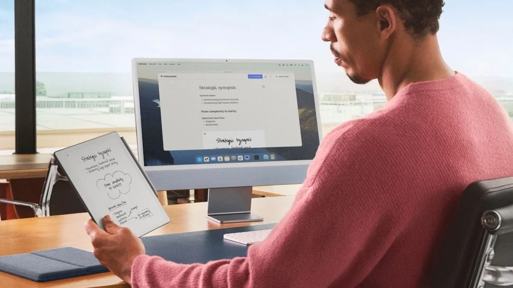 Person in a pink sweater using the reMarkable Paper Pure E Ink tablet at a desk beside a desktop monitor, with handwritten strategic notes syncing between the tablet and computer in a bright modern workspace