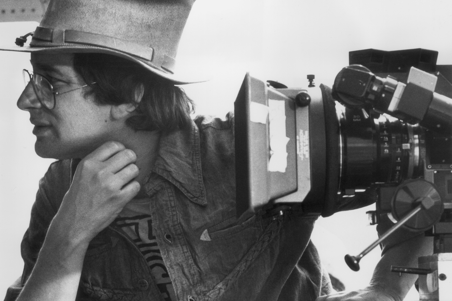 Black-and-white behind-the-scenes portrait of a filmmaker wearing glasses and a wide-brim hat while leaning beside a large motion picture camera setup during production