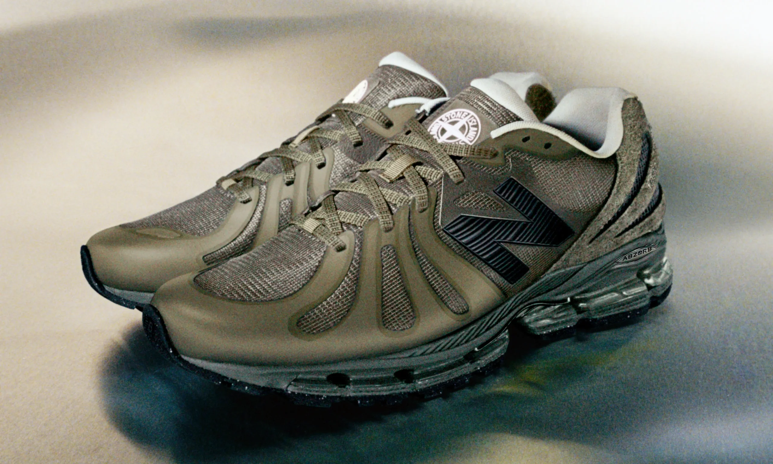Stone Island x New Balance ABZORB 1890 shoe in an earthy olive-grey colorway featuring technical ripstop mesh, molded wave-cut overlays, oversized black New Balance branding, and a visible full-length ABZORB cushioning unit photographed in a softly lit studio setting