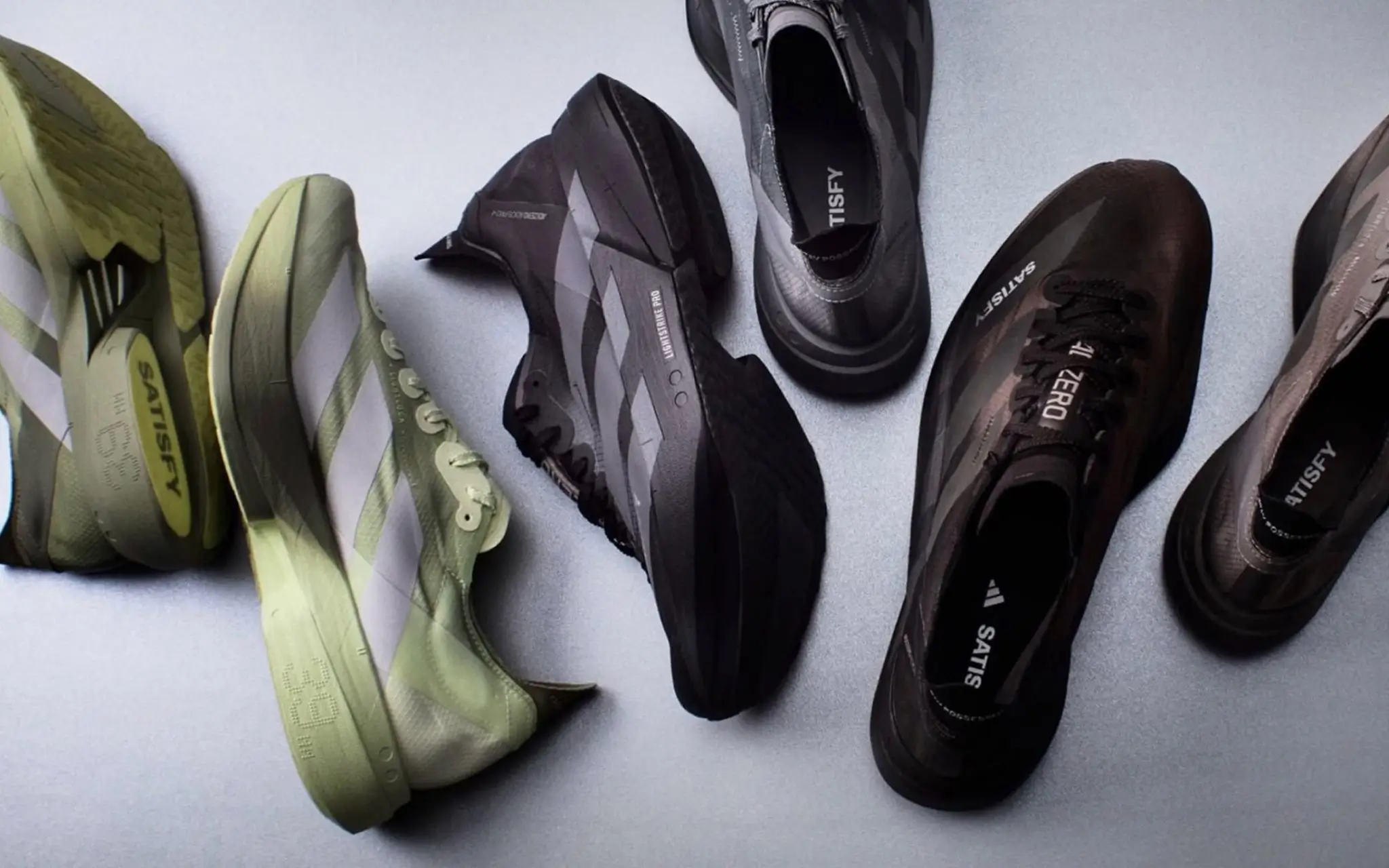SATISFY and adidas Adizero performance running shoes arranged in a minimalist overhead layout, showcasing lightweight race silhouettes in muted olive, black, and graphite colorways with aerodynamic midsoles and technical mesh uppers