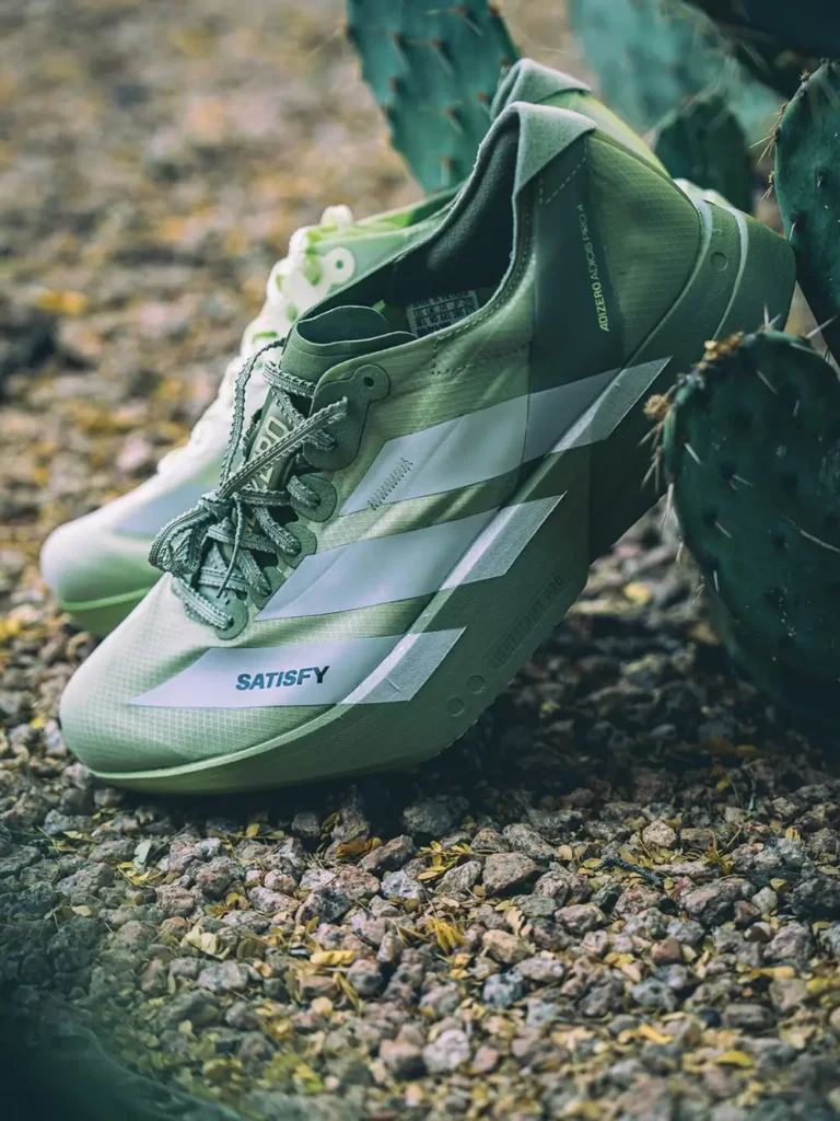 Olive green SATISFY x adidas Adizero Adios Pro 4 running shoes photographed outdoors beside cactus plants, highlighting lightweight mesh construction, oversized Three Stripes branding, and rugged desert-inspired performance style