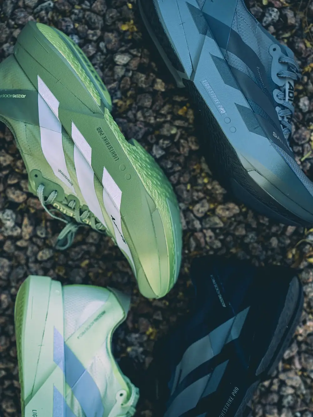 Multiple SATISFY x adidas Adizero Adios Pro 4 shoe displayed on rocky terrain in olive, white, grey, and black colorways, highlighting LIGHTSTRIKE PRO cushioning, angular Three Stripes detailing, and trail-inspired show design language