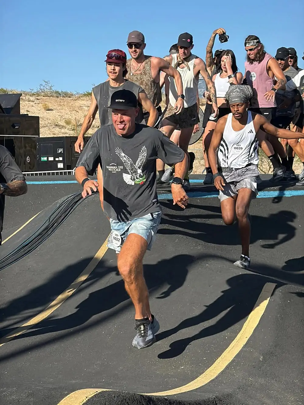 Runners sprint through a desert-inspired outdoor course during a SATISFY running event, wearing lightweight performance apparel and technical race footwear under bright afternoon sunlight