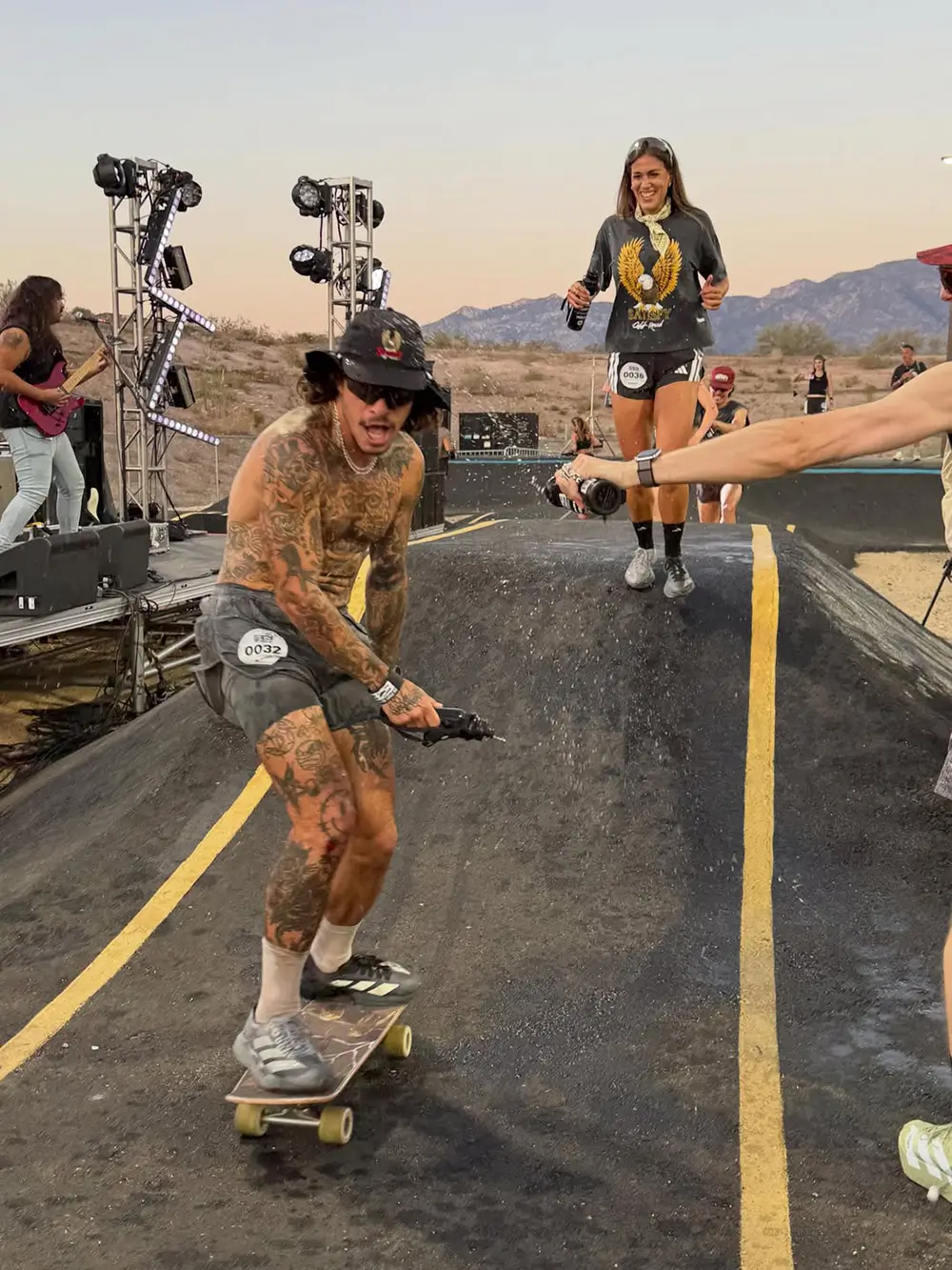 Tattooed skateboarder rides through a SATISFY outdoor running and music event at sunset, surrounded by runners, live stage equipment, and desert mountain scenery while wearing adidas performance footwear
