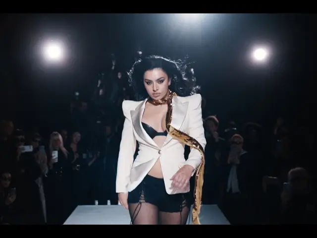 Runway scene from the “SS26” music video featuring Charli XCX walking toward the camera in a structured ivory blazer layered over black lingerie-inspired styling, finished with a dramatic metallic accessory and framed by intense show lighting and a fashion-week audience in the background