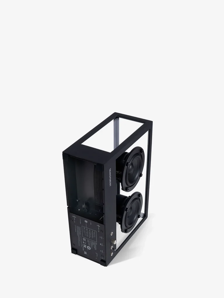 Angled top-down view of the Transparent Sound Small Speaker in Black featuring dual exposed drivers, transparent glass side panels, visible internal hardware, and a minimalist black aluminum frame against a light gray studio background