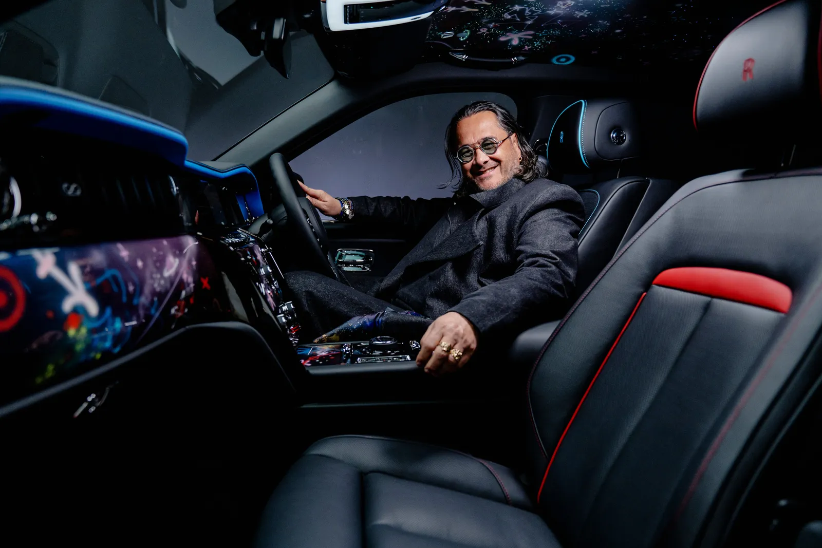Stylish designer seated inside a bespoke Rolls-Royce cabin illuminated by blue ambient lighting, showcasing cosmic-inspired dashboard artwork, celestial starlight headliner graphics, and black leather seats accented with vivid red detailing