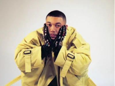 A portrait of a seated musician wearing an oversized beige technical coat with exaggerated proportions and extended straps pooling across the floor. The subject rests their face in black embellished gloves decorated with reflective jewel-like details, looking directly toward the camera with a calm, introspective expression. The minimal studio backdrop and muted palette place emphasis on silhouette, texture, and the contrast between utilitarian outerwear and expressive styling, creating a cinematic, fashion-editorial mood