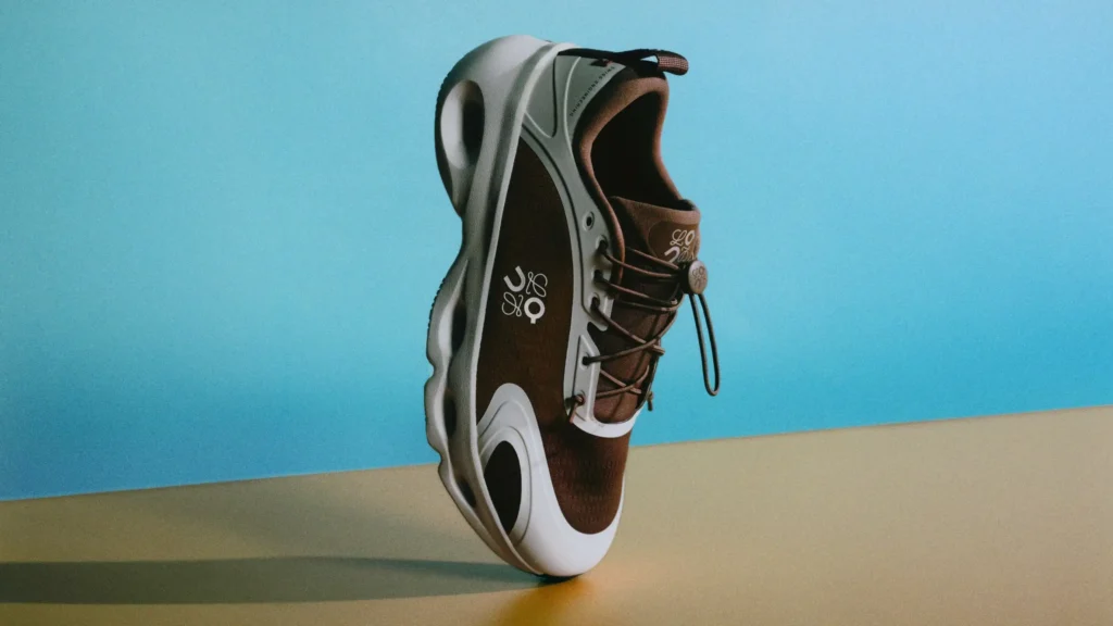 Editorial product image of a brown and cream Loewe performance shoe balancing upright on its sculpted sole, featuring oversized cutout cushioning, technical toggle lacing, and reflective logo detailing against a soft sky blue studio backdrop