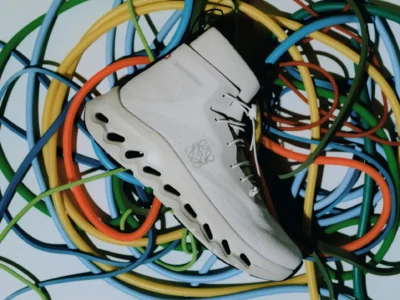 Editorial product image of a white Loewe high-top performance shoe with elongated hollow sole cushioning, placed atop a chaotic arrangement of multicolored cables under sharp studio lighting that emphasizes its futuristic knit construction and sculptural silhouette