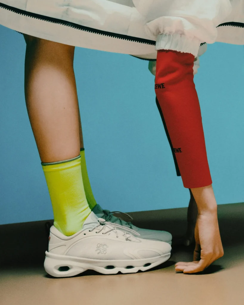 Close-up editorial image of a model balancing on fingertips beside oversized white Loewe slip-on sneakers with sculpted bubble soles, styled with neon yellow and crimson compression-style socks beneath a white technical garment against a powder blue backdrop