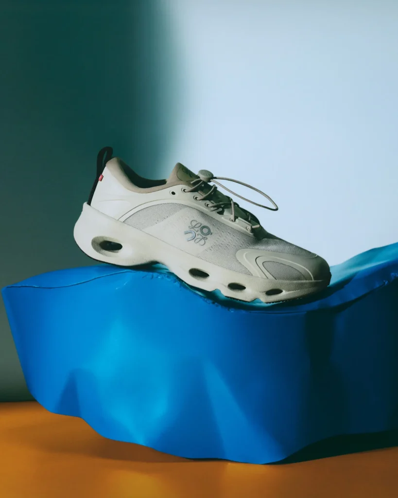 Editorial product image of a cream-toned Loewe shoe with sculpted perforated sole detailing, resting atop a fluid cobalt blue pedestal under dramatic studio lighting with soft shadow gradients and a muted grey backdrop