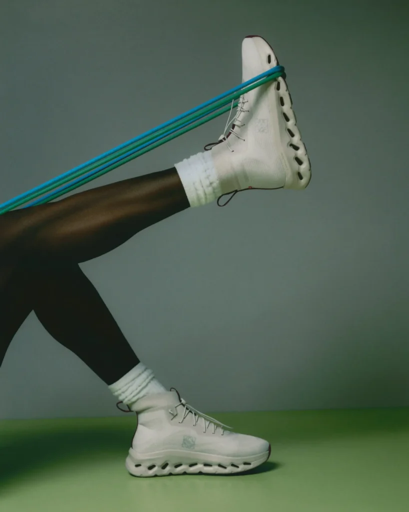 Editorial studio image of a model’s legs in motion wearing white Loewe high-top shoe with hollow sculpted soles, paired with ribbed white socks and stretched resistance bands against a muted green gradient backdrop