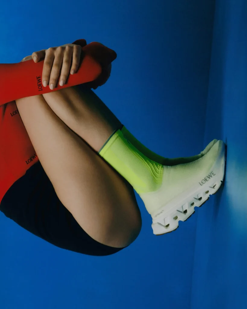 Editorial fashion image of a model curled against a vivid cobalt blue backdrop, wearing neon green Loewe show socks and white sculpted trail-style sneakers paired with bold crimson arm sleeves featuring subtle Loewe branding