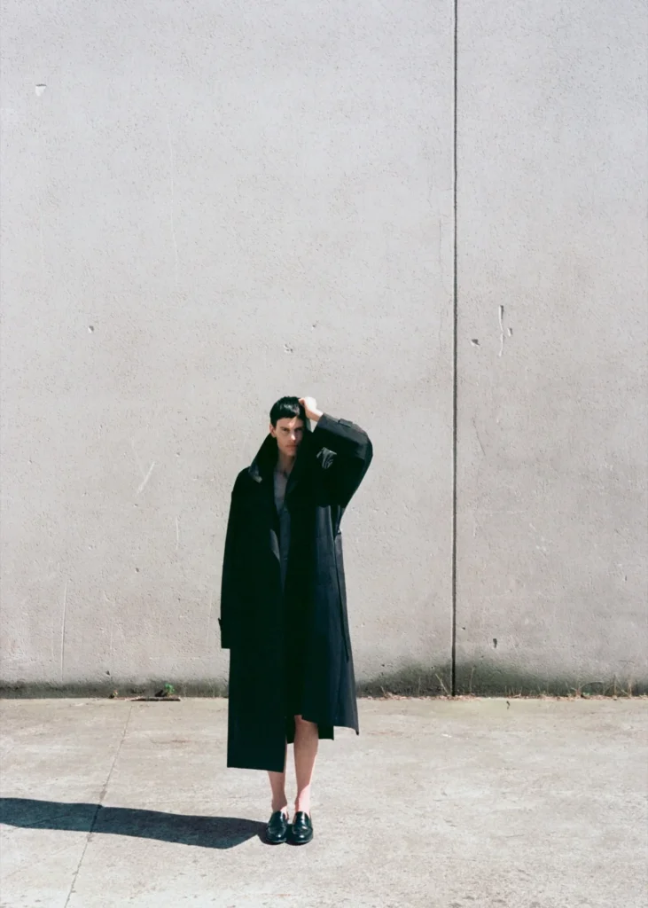 Model standing against a large concrete wall wearing an oversized black ssstein overcoat with a dramatic collar and black loafers, photographed in bright natural sunlight with long shadows and minimalist architectural surroundings