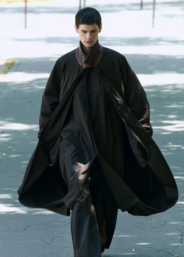 Model walking outdoors in a flowing oversized black ssstein coat with a sculptural brown leather collar and fluid dark trousers, photographed in soft daylight with silhouette cast across the pavement