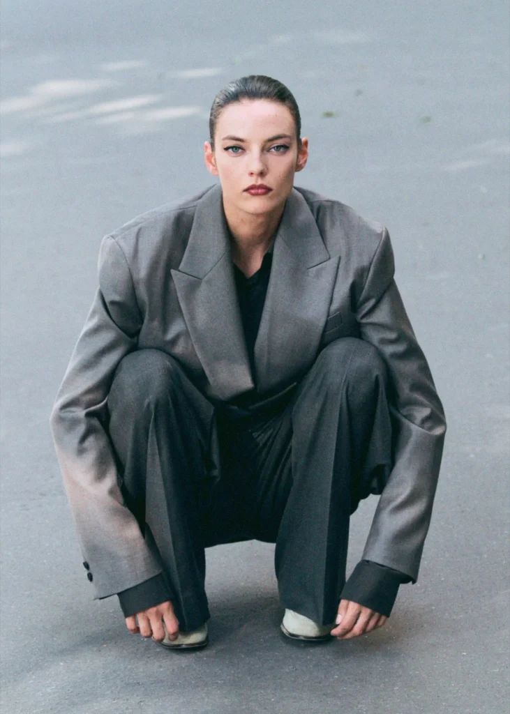 Model crouching on pavement wearing an oversized charcoal ssstein tailored suit with broad shoulders, fluid trousers, and a minimalist monochrome aesthetic